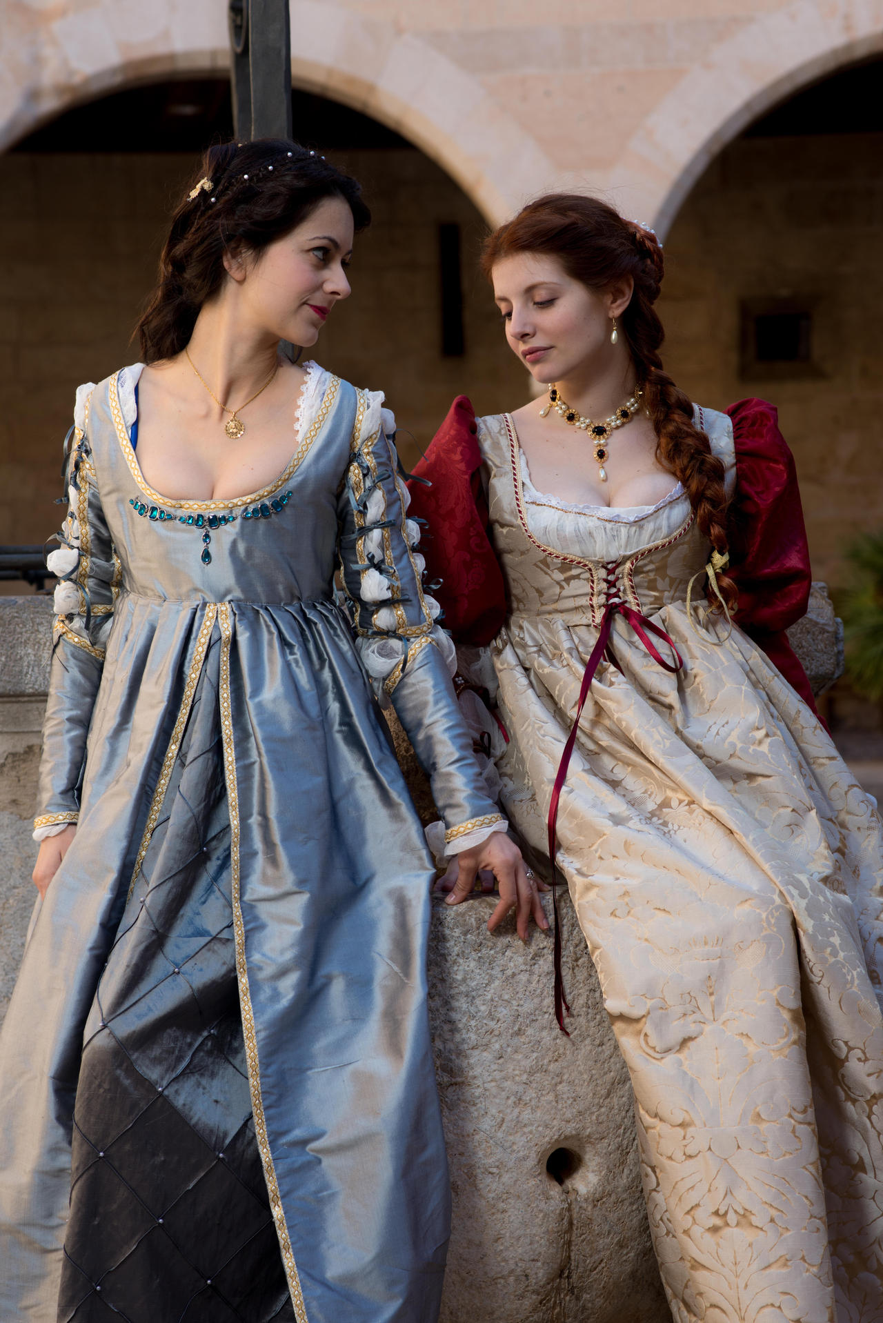 two women, dressed in elaborate renaissanceera gowns, stand together in a stone courtyard.  the woman on the left wears a light blue gown with gold detailing, while the woman on the right wears a creamcolored gown with burgundy sleeves.  they appear to be engaged in conversation. the setting suggests a historical reenactment or a scene from a historical film. the detail in the clothing is exquisite, showcasing the craftsmanship and artistry of the period.