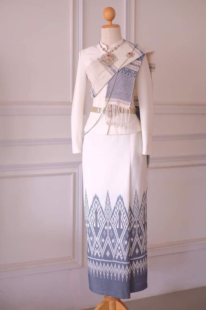 a closeup of a white thai dress with a blue patterned border. the dress is worn on a mannequin and is decorated with a silver belt, a silver pendant necklace, and a blue and white silk shawl. the mannequin is standing in front of a white wall with decorative white moldings.  the dress is traditional thai clothing for women.