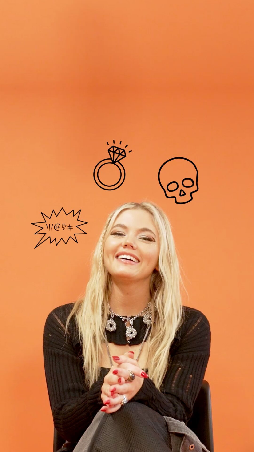 a blonde woman with a black top and layered necklaces is sitting against an orange background with doodles of a ring, skull, and profanity symbol above her head. she is smiling and has her hands clasped together. she is wearing multiple rings.