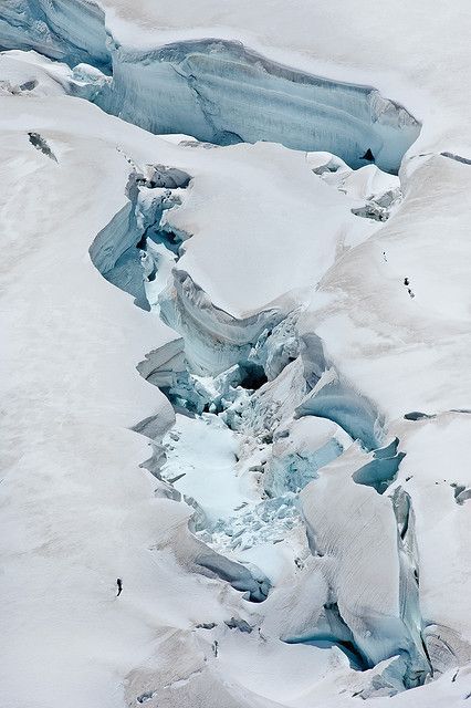 aerial view of a vast snowy landscape featuring a deep crevasse cutting through the ice. the crevasse reveals a blueish ice beneath the snow, creating a dramatic contrast. a solitary person, appearing small in comparison to the expansive scene, stands near the edge of the crevasse, likely a mountaineer. the environment is cold and icy, suggesting a highaltitude or polar region.