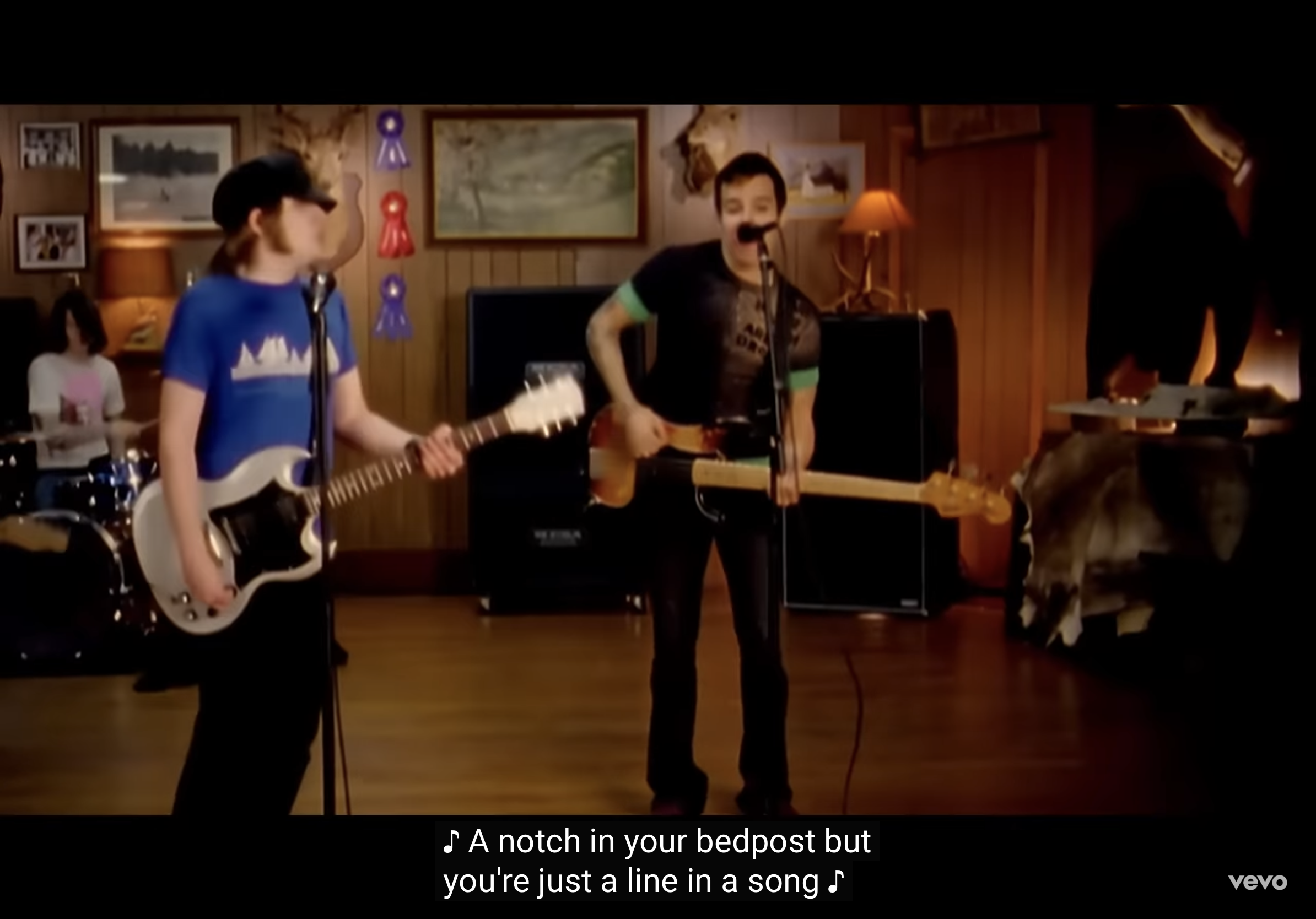 a still from what appears to be a 2000s rock music video. four band members are playing in a room with woodpaneled walls. the singer is center right, holding a microphone and wearing a black graphic tshirt. a female guitarist in a blue tshirt stands to his left, and a drummer and bassist are visible in the background.  the lighting is warm and artificial. the lyrics "a notch in your bedpost but you're just a line in a song" are displayed at the bottom of the image.