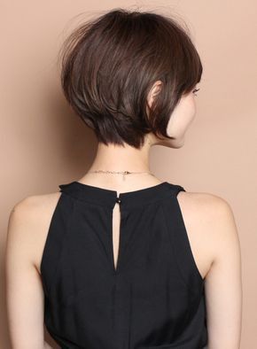 a woman with short brunette bob with bangs is looking over her shoulder. she is wearing a black sleeveless top. her hair is styled in a way that shows off her neck, which has a gold necklace. the image is taken from the back, giving a good view of the hair style.
