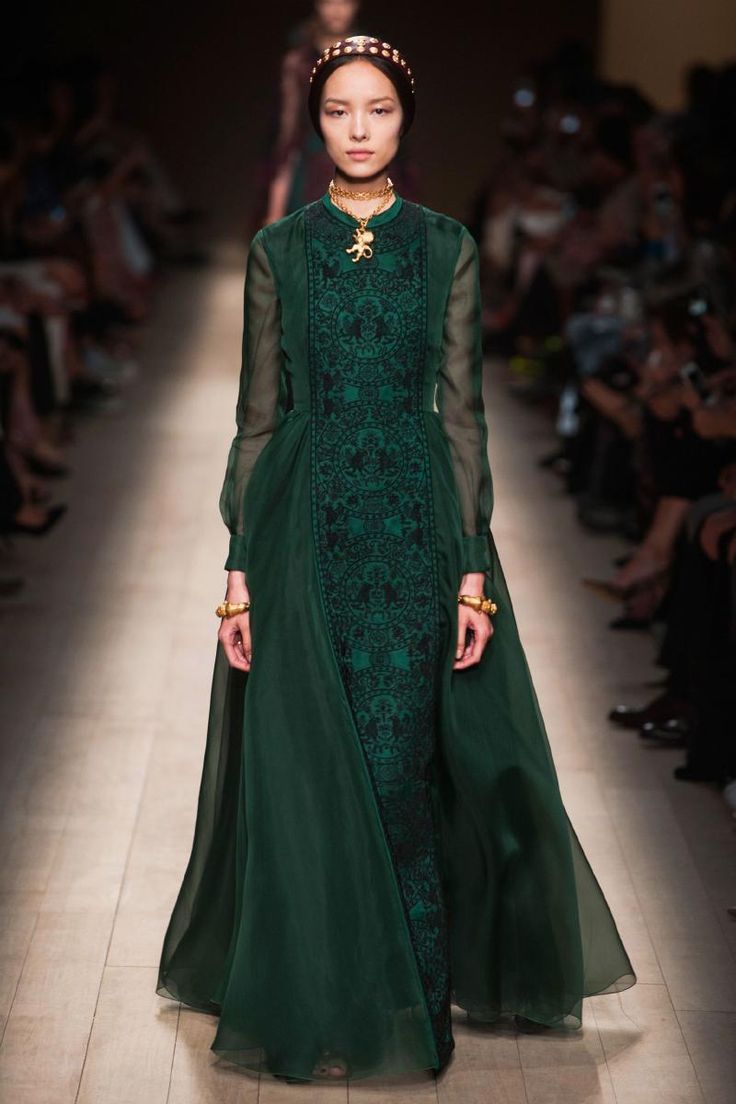 a model walks down a runway wearing a long, elegant emerald green gown. the dress features an embroidered bodice and a sheer overlay. the model is accessorized with a gold headband, necklace, and bracelets. the overall style is sophisticated and high fashion.  the background is blurred, showcasing other models and audience members. the setting is clearly a professional fashion show.