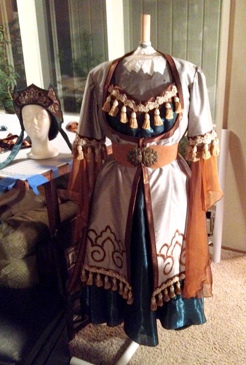 a closeup shot of an elaborate costume, possibly for a theatrical performance or historical reenactment, displayed on a mannequin. the dress is predominantly offwhite with teal and brown accents.  it features intricate embroidery, gold embellishments, and numerous tassels. a matching headpiece sits on a separate mannequin nearby. the setting appears to be a home or workshop, with a table and other items visible in the background. the overall aesthetic suggests a handcrafted, detailed, and highquality garment.