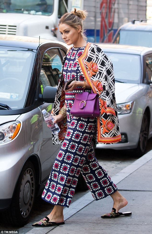 the image shows olivia palermo, a known fashion icon, on a city street.  she's wearing a stylish and colorful outfit. this includes a dark navy blue and red crocheted jumpsuit with wideleg palazzo pants. over this, she wears a black and white embroidered jacket with orange accents. she carries a bright purple  handbag (possibly dolce & gabbana) and is holding a water bottle. she's wearing dark embroidered mules.  the background includes parked cars and urban elements. the overall impression is one of highfashion street style.