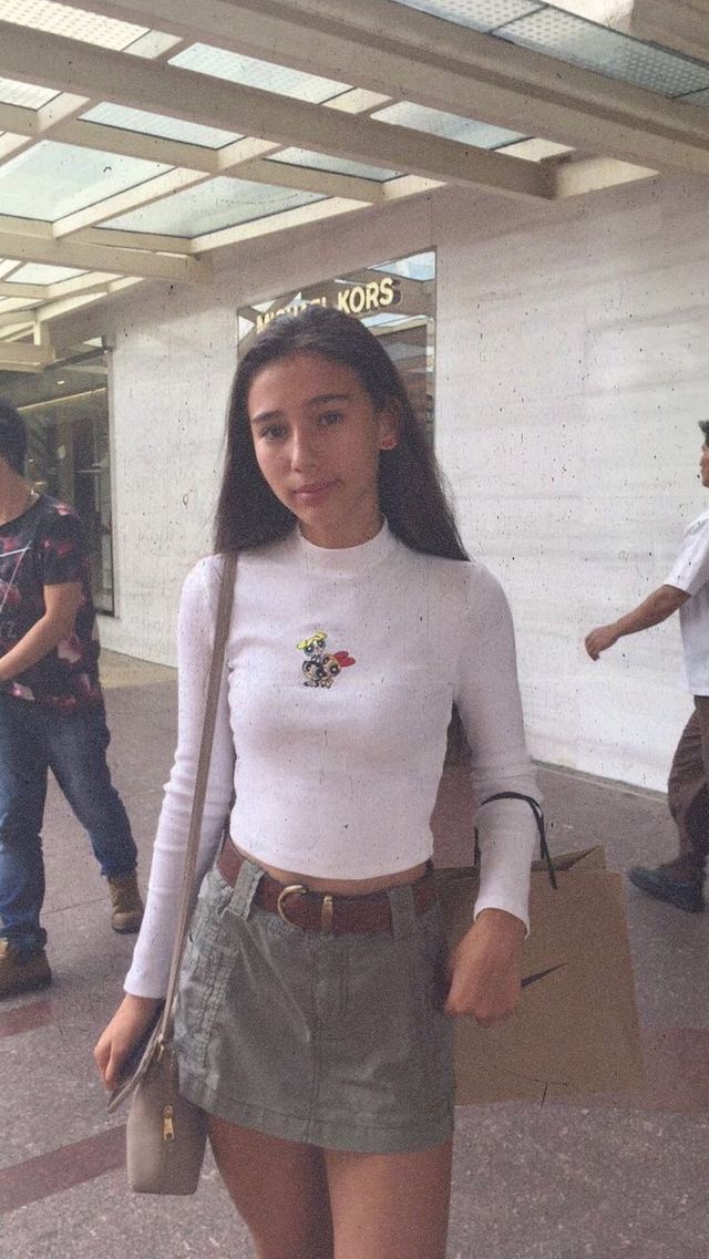 a young woman is standing outside a store. she has long dark hair and is wearing a white long sleeve tshirt, a short khaki green skirt with a brown belt, and carrying a purse.