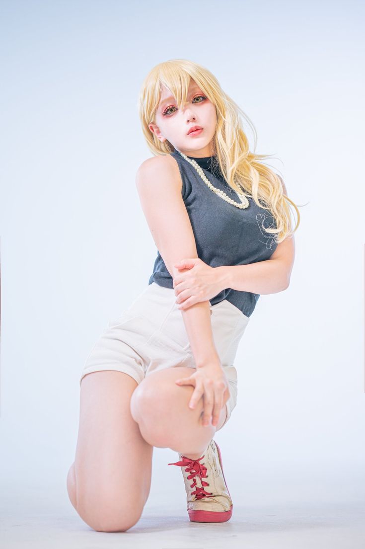 a young woman with long blonde hair is cosplaying in a studio setting. she is kneeling with her body angled slightly toward the camera. she is wearing a black sleeveless top, white shorts, and offwhite sneakers with red laces.  she also wears a pearl necklace.  her makeup is clean and emphasizes her eyes. the background is a plain white backdrop.  the overall image is clean, bright and well lit.