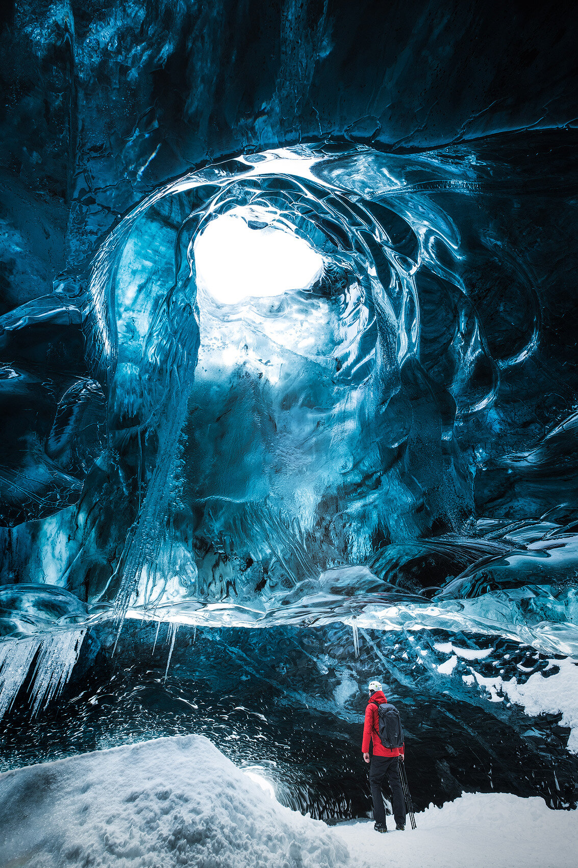 a lone figure stands in awe at the entrance of a stunning blue ice cave, the light filtering through the icy ceiling casting an ethereal glow. the vastness and beauty of the ice formations are evident, creating a magical and otherworldly scene.