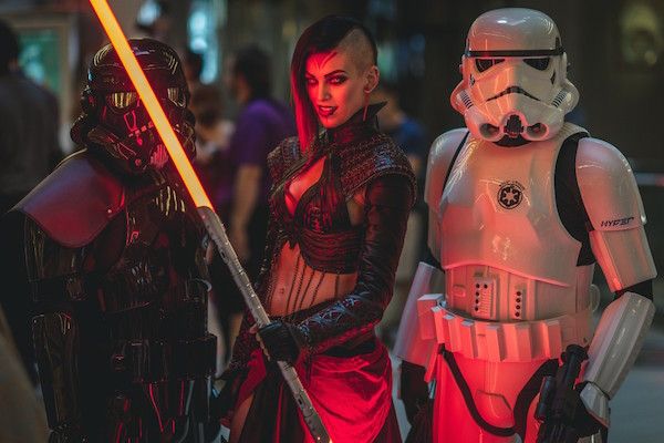 a woman in a red and black costume with a lightsaber, a stormtrooper, and a dark figure with a lightsaber are seen in an action pose.  the woman has a shaved head with red streaks in her hair and is holding a glowing orange lightsaber.  the stormtrooper is in a classic white and black costume and is standing in a defensive stance with a blaster in hand.  the dark figure is wearing black armor and is holding a glowing orange lightsaber.  the scene is set against a dark background with some lighting effects.