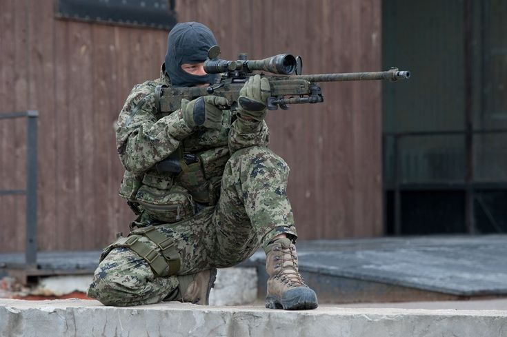 a soldier in camouflage gear is aiming a scoped rifle. the soldier is wearing a black balaclava and has a serious expression on his face. he is kneeling on a concrete surface in front of a wooden building. the soldier is likely a sniper and is prepared for combat.