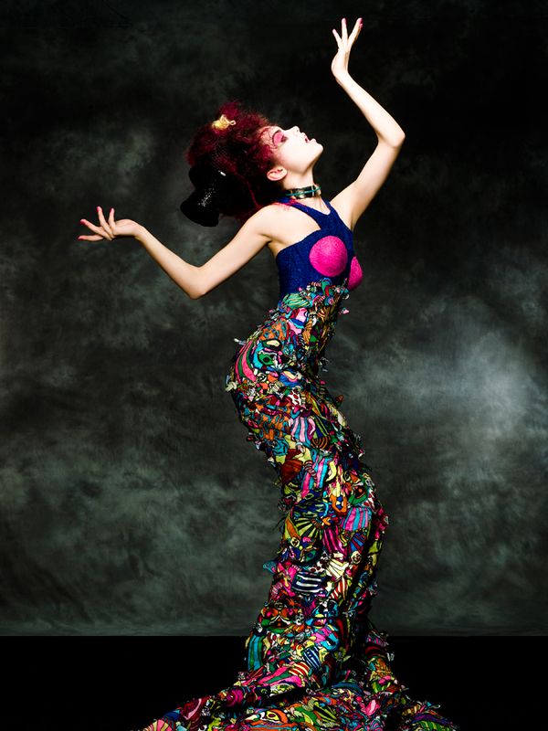 a woman in a brightly colored dress is posing dramatically in a studio. her arms are stretched out and she is looking up, her red hair is styled in a dramatic fashion.