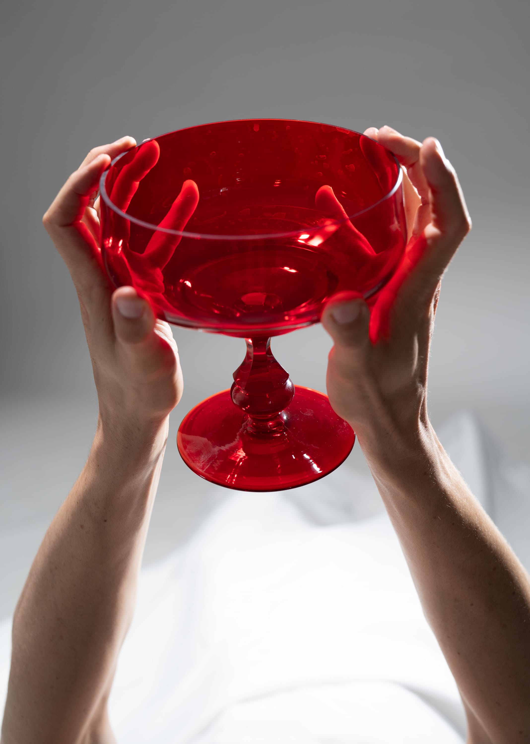 a woman is holding up a red glass bowl with a pedestal base against a white background. the image is lit with soft studio lighting.