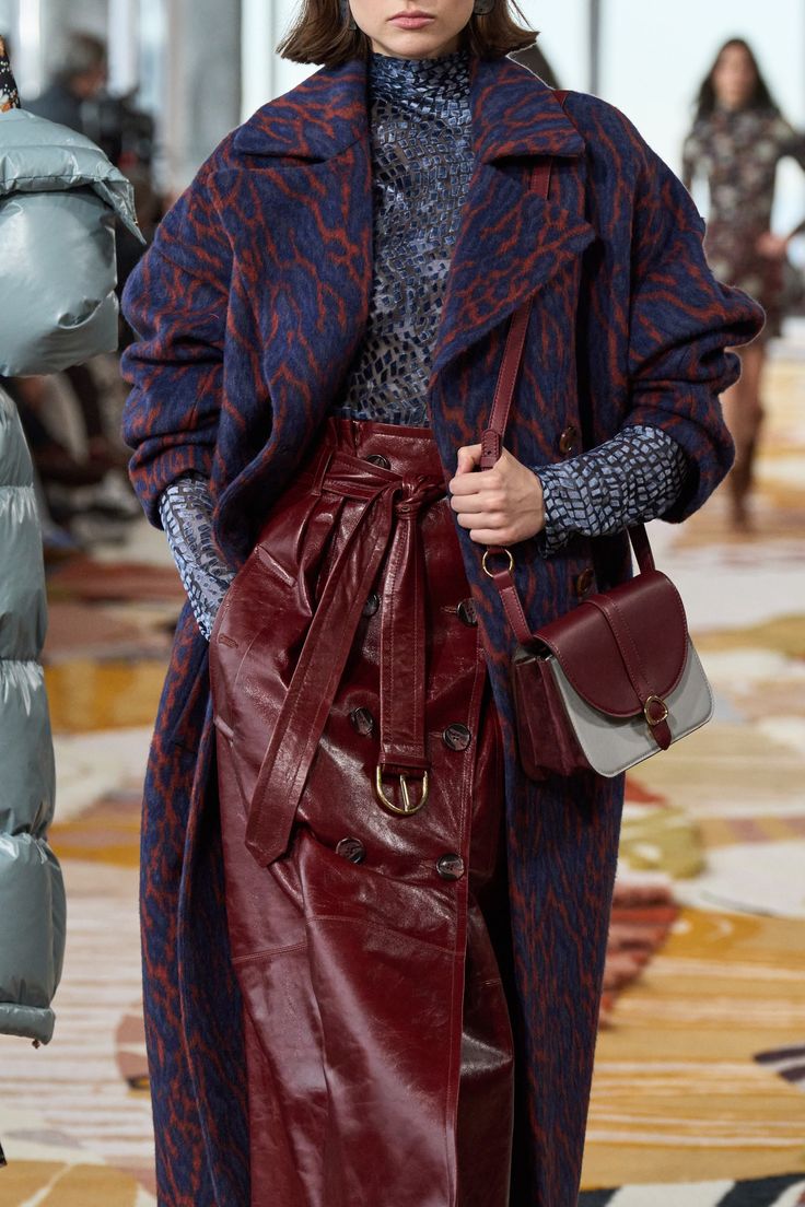a woman is showcasing a stylish ensemble on a runway. she's wearing a dark blue and brown leopardprint coat paired with a rich burgundy leather skirt. the skirt features a belted waist and button detailing. completing the look is a crossbody handbag in a combination of burgundy and grey.  the model's turtleneck is visible beneath the coat, adding layers to the outfit. the background subtly suggests a fashion show setting.