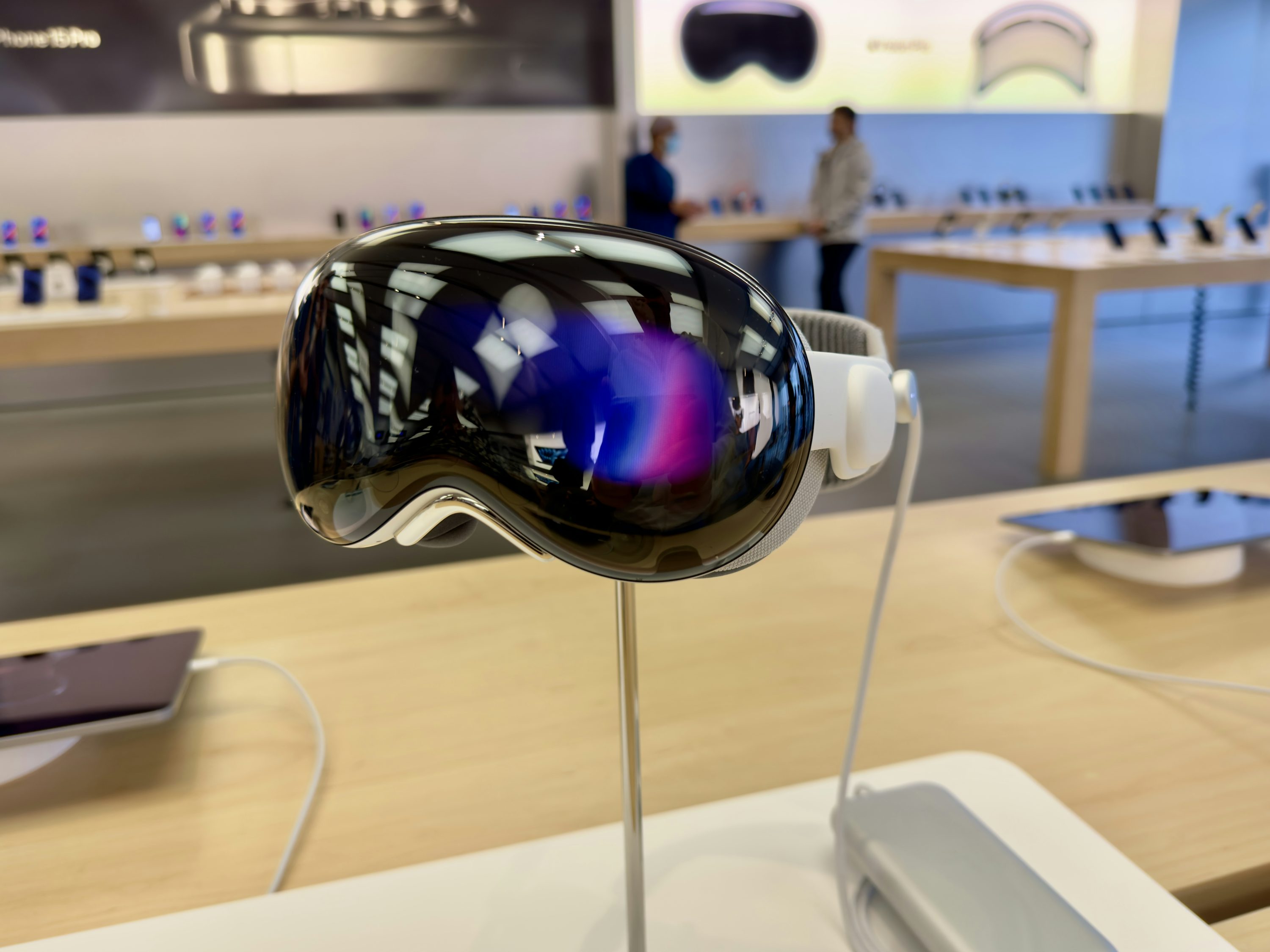 a closeup shot of an apple vision pro vr headset on display in an apple store. the headset is mounted on a chrome stand and has a shiny, reflective surface. an ipad on a wireless charger is sitting next to the headset. other apple devices and customers can be seen blurred in the background.