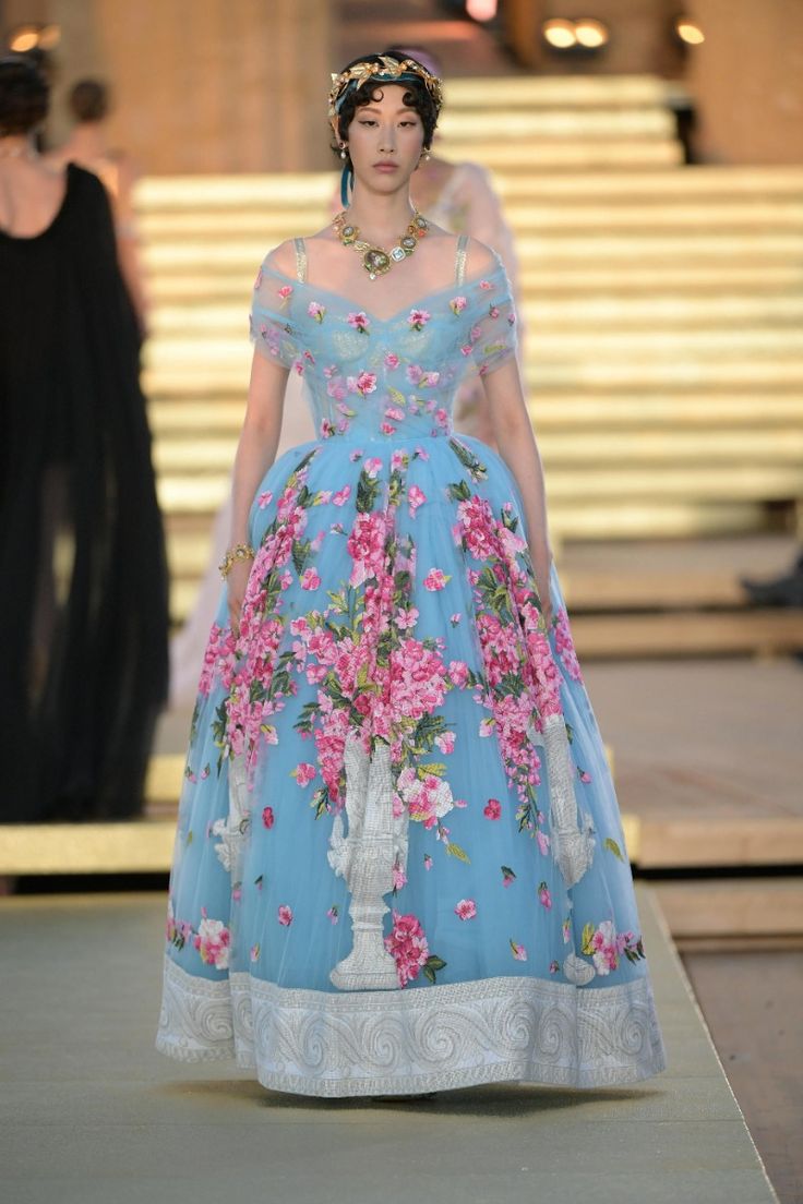 a model walks the runway wearing a stunning blue gown with pink floral print. the gown is fulllength and has a fitted bodice and a flowing skirt. the model is wearing gold jewelry, including a necklace and earrings. she has a headband with a gold accent in her short, dark hair, and a dramatic eye look with bold eyeliner and bright lipstick. the backdrop of the runway is out of focus, with a stone wall and stairs visible.  the model is posed with her head held high and a confident expression, showing off the intricate details of the gown and the luxurious look. the image is a snapshot of high fashion, showcasing the beauty and artistry of couture design.