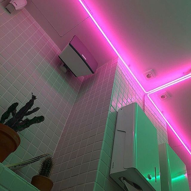 a modern bathroom with pink neon lights illuminating the white tiled walls and a toilet paper holder. the tiles are a light gray and the paper holder is a sleek white design. there is a cactus plant on a shelf below the paper holder.  the neon lights add a touch of modern design and create a cool and sophisticated atmosphere.