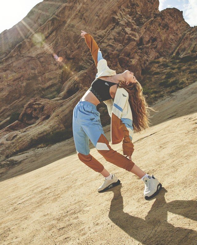 a young woman is posing in a desert setting. she is wearing a brown jacket, a blue and white athletic outfit, and white reebok sneakers. she is striking a pose with one arm raised and looking upwards, as if she is about to take off. the sunlight is bright, and the mountains are visible in the background.  she looks confident and ready for action.  this image is a good representation of a stylish, athletic woman who is living a healthy and active lifestyle.