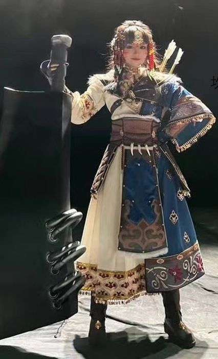 a woman in a fantasy warrior cosplay costume is standing and looking at the camera. she is holding a sword in her left hand. she is wearing a blue and white dress with gold trim, and her hair is in braids. she is wearing a brown belt with a buckle and a bow and arrow are on her back. the background is dark, and the image is lit from the front.