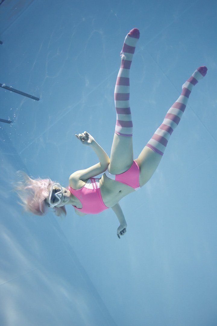 a young woman with pink hair is captured swimming underwater in a pool. she is wearing a pink and white striped bikini top and bottom, long pink and white striped socks, and pink swimming goggles. the perspective is from below, looking up towards the bright, sunlit surface of the water.  the image conveys a sense of freedom, joy, and summer fun.