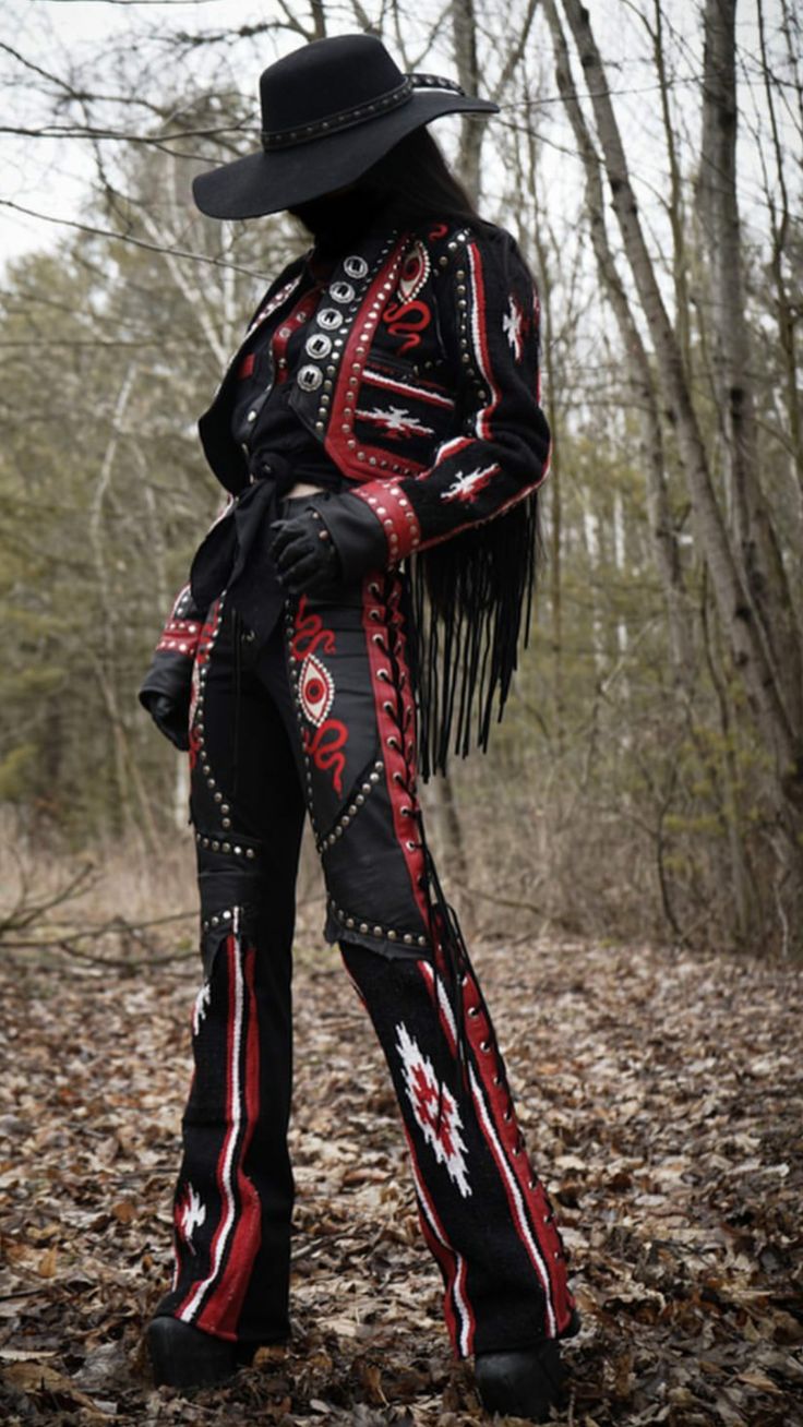 a woman wearing a stylish cowboy hat, a black jacket with red and white embroidery, and black pants with a red and white patterned trim stands in a forest with fallen leaves. she has a serious expression as she looks down. her jacket has fringe on the bottom and an embroidered snake and eye. the pants are lace up and also have eye embroidery.  she is wearing black boots and a black belt.