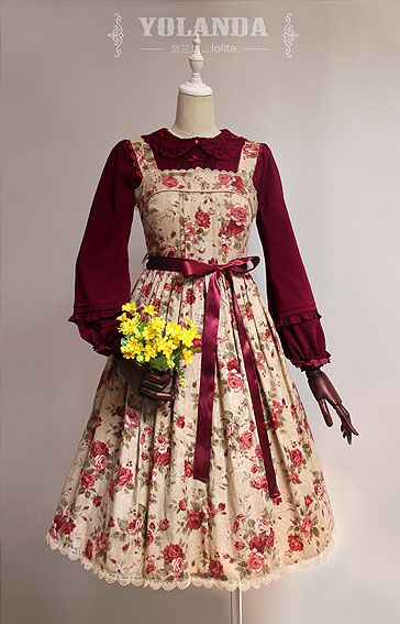 a creamcolored lolita dress with a pink and red floral pattern. it has a square neckline, long sleeves, and a burgundy velvet pinafore with a matching belt. it also has a peter pan collar with lace trim. the dress falls to the knee and is displayed on a mannequin against a plain background.