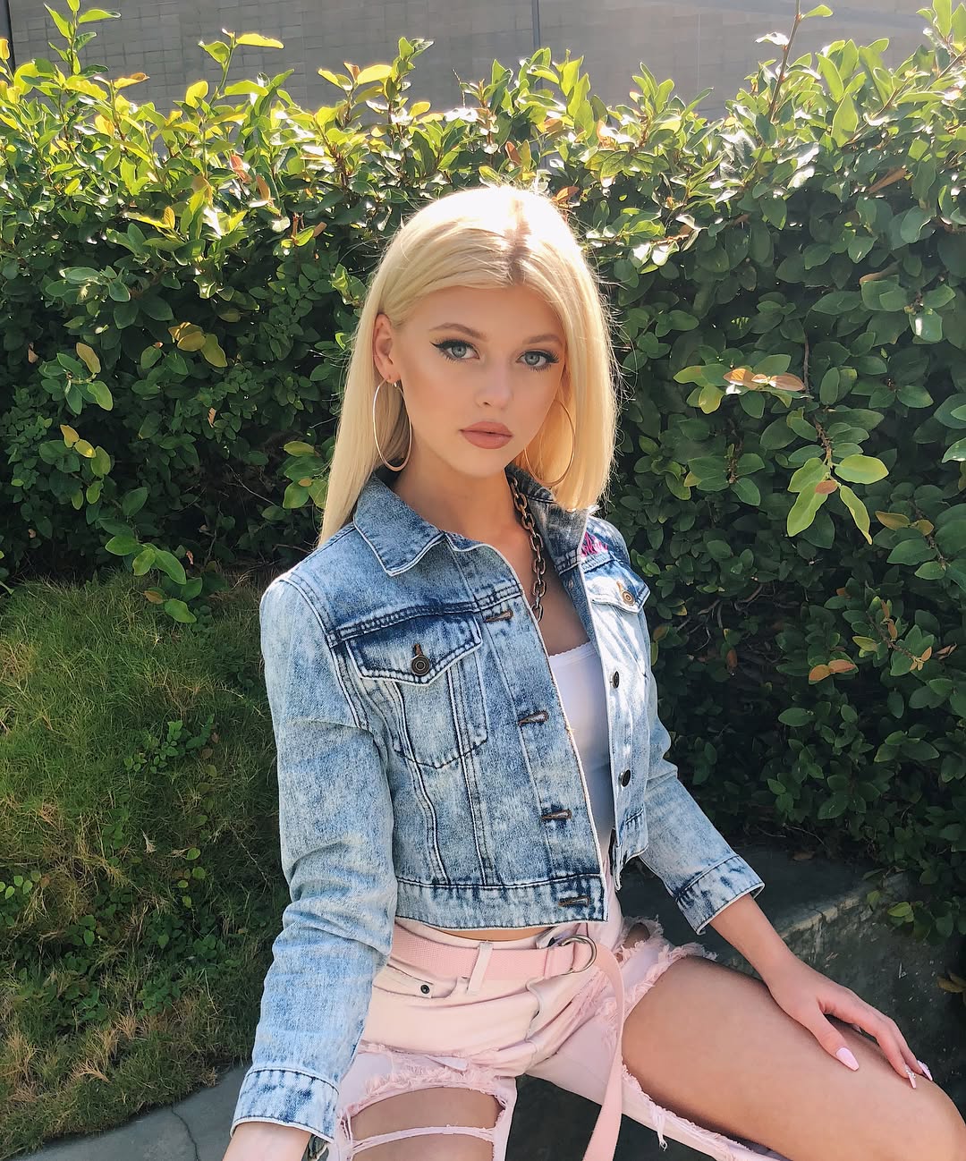 a blonde woman with hoop earrings, wearing a denim jacket and pink shorts, is sitting in a garden setting.