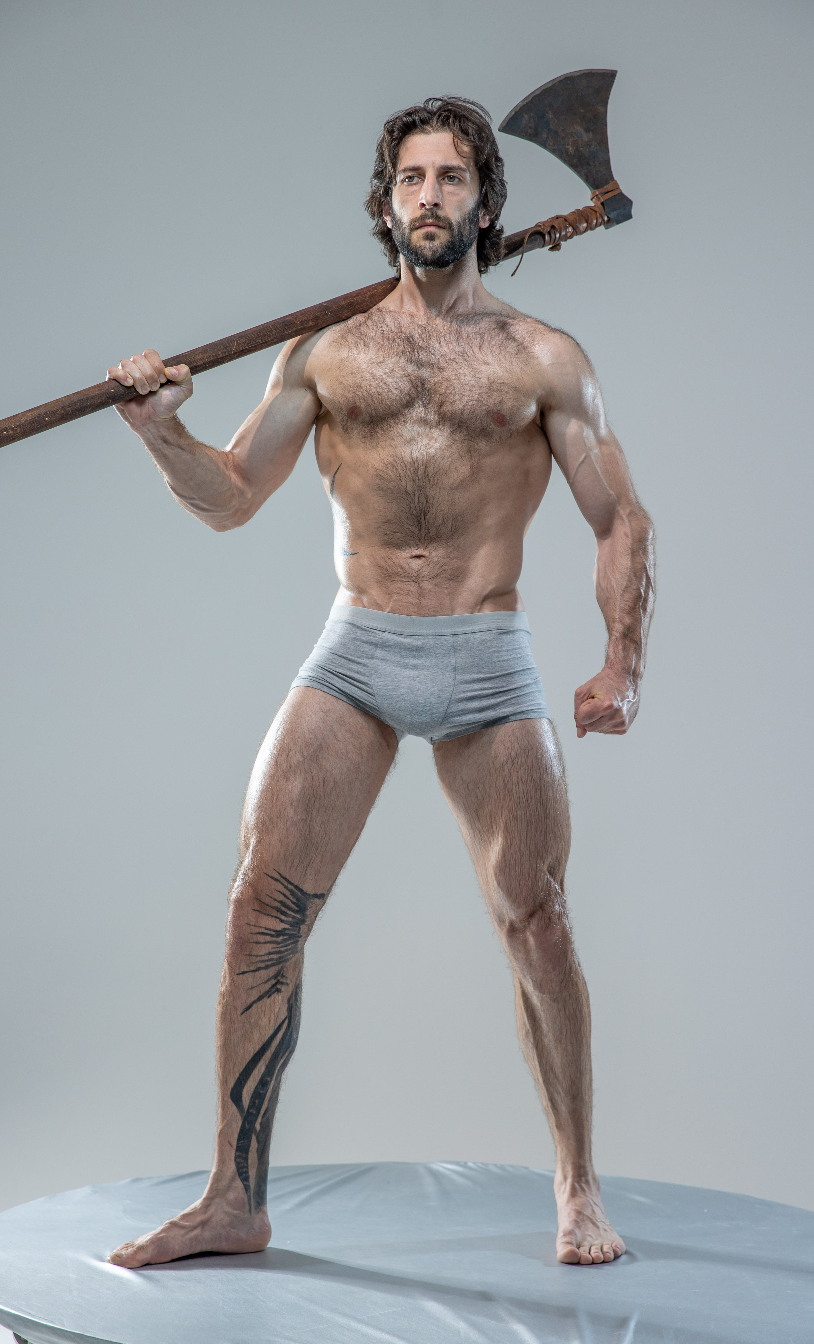 a muscular man with long hair and beard, stands shirtless, wearing only grey underwear, on a round platform against a light grey backdrop. he confidently holds a large, worn axe over his right shoulder, his left hand clenched in a fist. he has a tattoo on his left leg. the image exudes strength, power, and a hint of aggression.