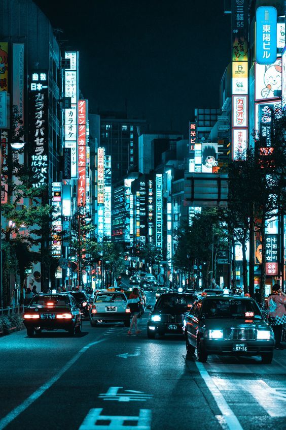 a blurred, nighttime photograph of a street in tokyo, japan. the street is illuminated by neon signs and car headlights. cars are driving down the road in both directions, and there are buildings lining both sides of the street. the image has a futuristic, cyberpunk aesthetic.