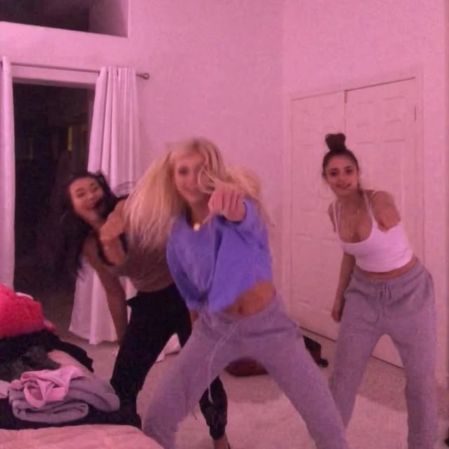 three women are posing for a picture indoors in what appears to be a bedroom. the woman in the center is wearing a purple longsleeve crop top and light gray sweatpants, pointing towards the camera. the woman on the left is wearing a brown top and black pants. the woman on the right is wearing a white tank top and light gray sweatpants. the room is decorated with a doorway and a curtain. there's a bed with clothes on the left side of the image. the overall image has a pinkish hue.
