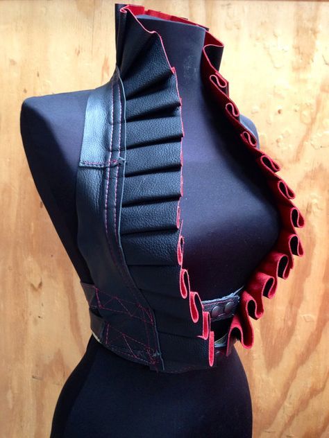 a black leather vest with red ruffles is displayed on a mannequin. the vest has a high, ruffled collar and a layered design on the front. it is fastened with a belt around the waist.