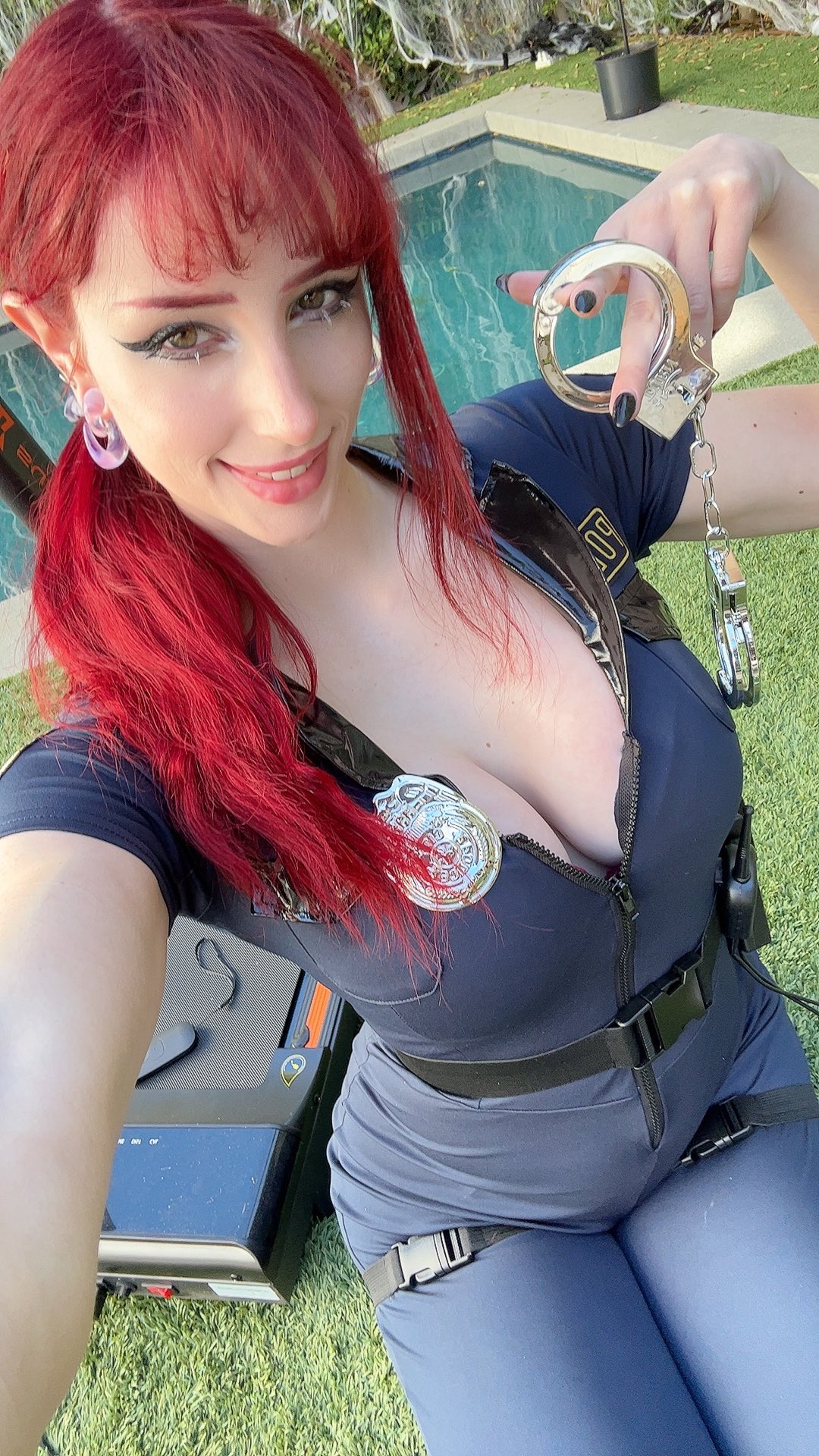 a woman with bright red hair, wearing a police officer costume, takes a playful selfie outdoors.  she's holding a pair of handcuffs and is smiling directly at the camera. the setting appears to be by a swimming pool with grass in the foreground. her costume is detailed, showing a badge and utility belt. the overall mood is flirty and fun.