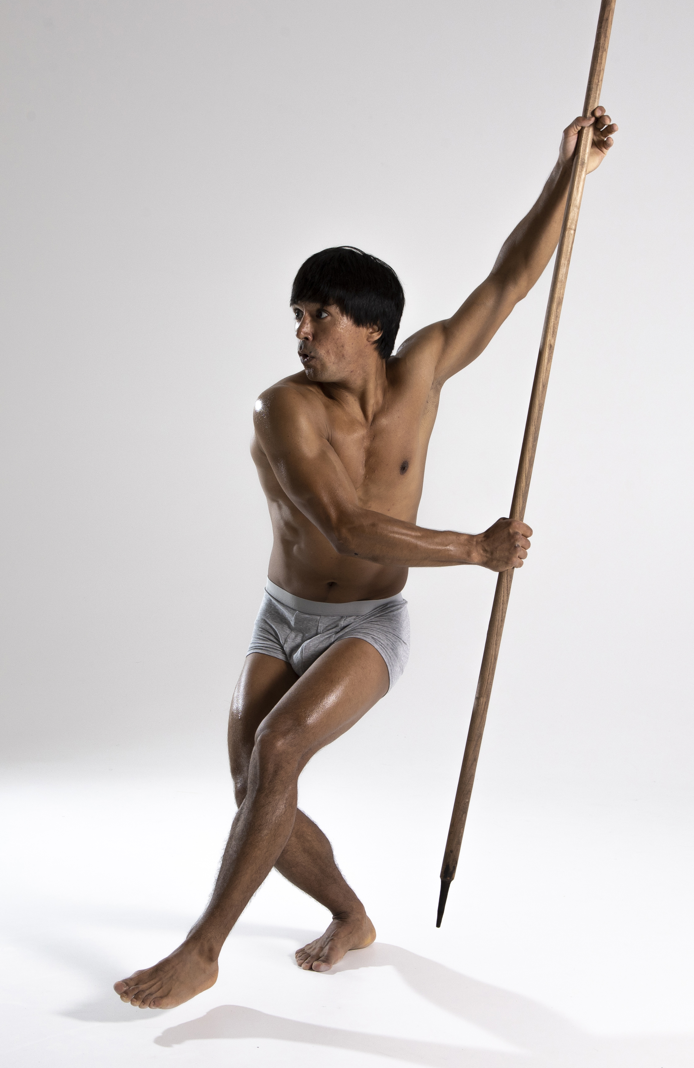 a shirtless man in underwear is posed against a light background, holding a spear.