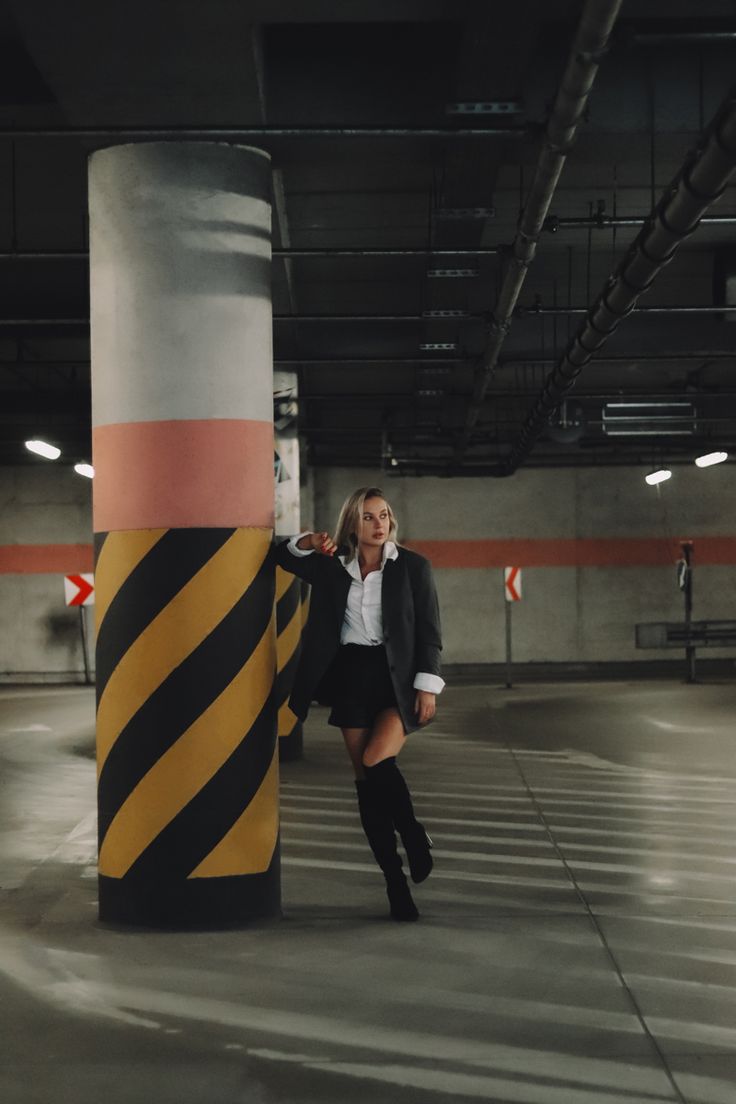 a young woman stands in a parking garage, leaning against a striped concrete pillar. she is wearing a black blazer, a white shirt, a black skirt, and kneehigh black boots. she is looking to her left, her blonde hair falling over her shoulders.  the lighting in the garage is dim, and the concrete floor is gray. the image is a bit dark, but the woman's outfit and pose are clear and the overall look is chic and sophisticated.