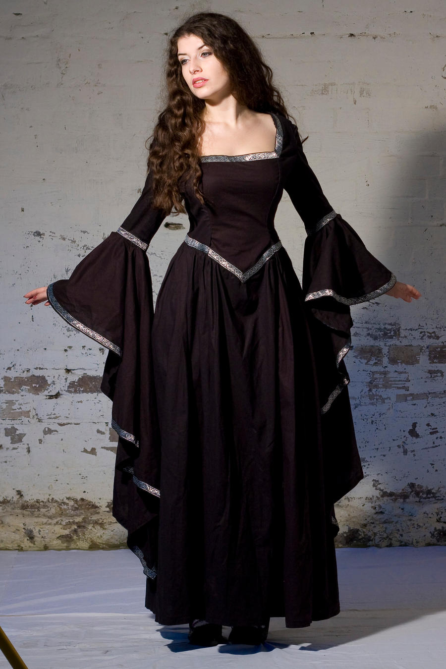 a fulllength shot of a young woman in a long, dark medievalstyle dress. the dress is black with silver accents at the neckline and sleeves, which are long and bellshaped. she has long, dark hair and is posing against a whitewashed brick wall. the overall style is gothic or fantasyinspired. the image is welllit and the details of the dress are clearly visible.