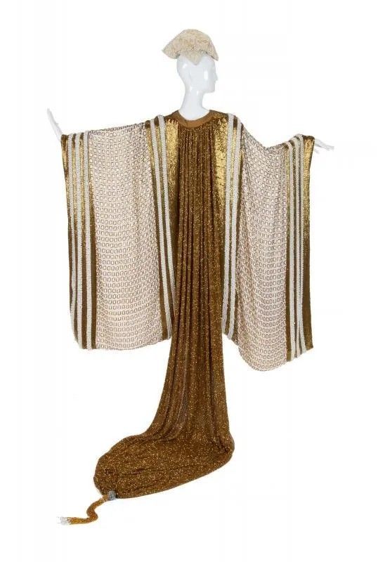 a mannequin wearing a gold and white dress with a matching headdress stands against a white background. the dress features a gold center panel that extends into a long, draped train with tassels, while the sides are white with an intricate pattern and accented with gold stripes. the headdress is made of the same fabric as the side panels of the dress.