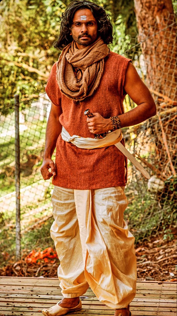 the image shows a man of south asian descent, possibly indian, dressed in traditional attire. he's wearing an orange kurta (a loosefitting shirt), a beige dhoti (a type of wrapped garment), and a brown scarf draped around his neck. a white sash is around his waist, and he holds a sword.  a tilak (a mark on the forehead) is visible, suggesting a religious or spiritual context. he has long dark hair, and his expression is serious. the setting appears to be outdoors, with a wooden background and some foliage visible. the overall style and attire suggest a representation of a hindu character, potentially a warrior or religious figure.