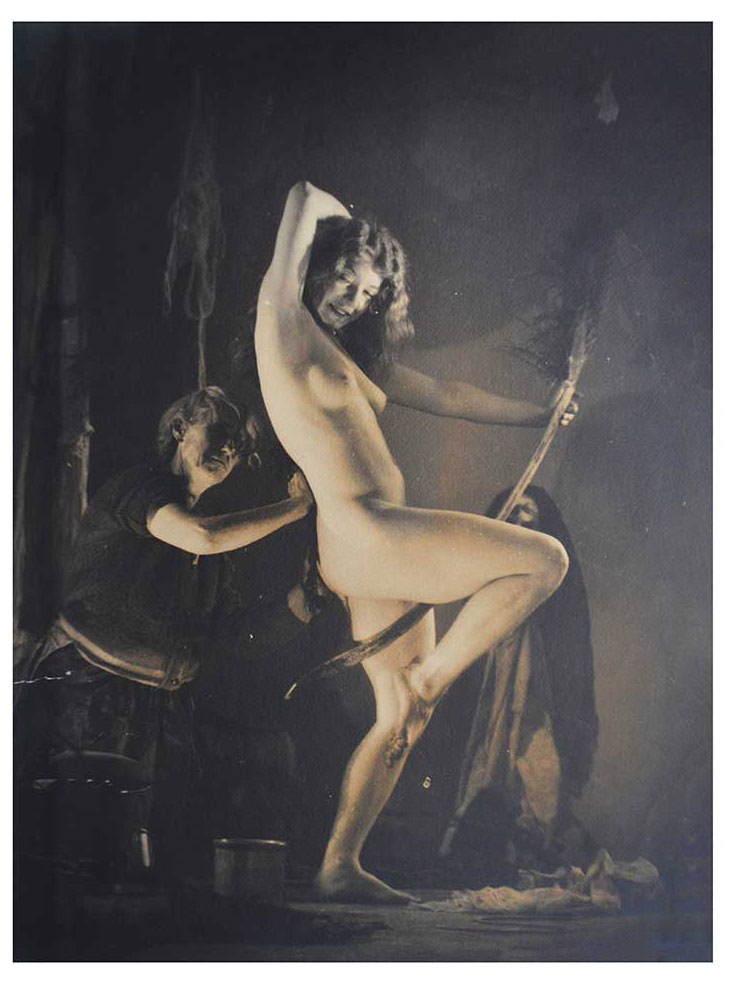 the image is a vintage sepiatoned photograph, likely from the early to mid20th century, depicting a nude young woman as the central figure. she is posed dynamically, her body arched, holding a long staff or broomlike object. the lighting is dramatic, creating deep shadows and highlighting her form. two older women, seemingly assisting or observing, are partially visible in the background, suggesting a ritualistic or ceremonial context. the overall mood is mysterious, sensual, and somewhat unsettling. the style suggests an artistic approach influenced by pictorialism, a movement in photography emphasizing artistic expression and manipulation of the image.  the dark background and muted tones add to the somber atmosphere.