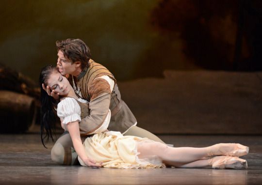 two dancers in mid performance. the woman is in a white and creamcolored dress and the man is in a  brown leather jacket.  they are in a relaxed position on stage.  the man is holding the woman's hand and leaning toward her.  they are looking at each other with affection.