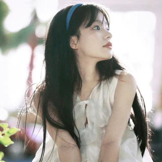 a young woman with long black hair wearing a white dress and a blue headband is sitting outdoors looking away from the camera. she has a dreamy and youthful appearance. the lighting is natural and bright.