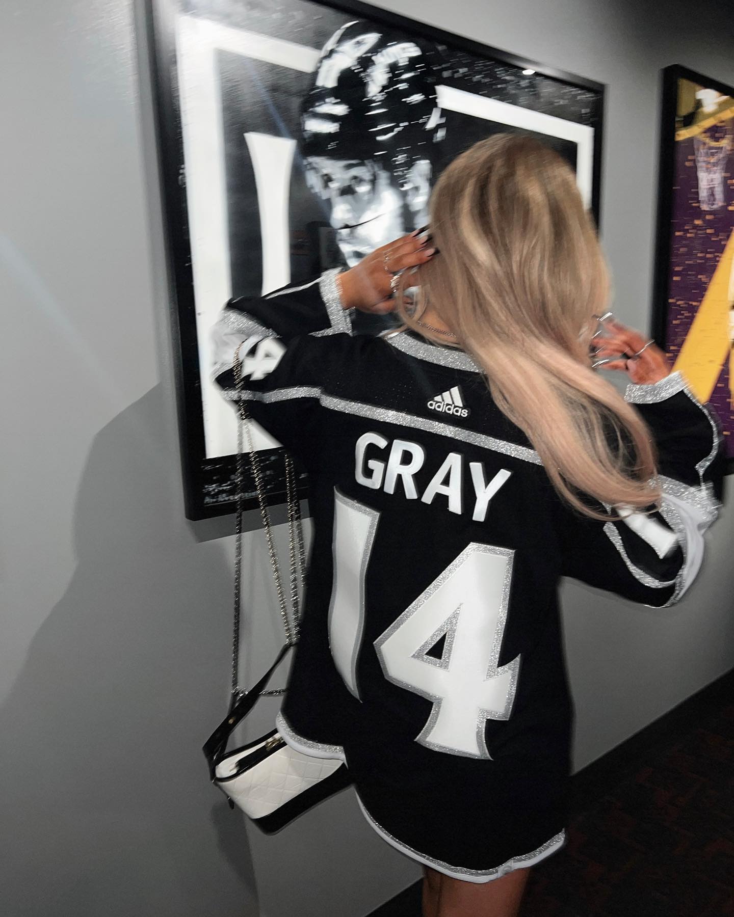 a woman with blonde hair is seen from the back, wearing a black and white hockey jersey with "gray" printed on the back along with the number "14". she has a purse with a chain strap hanging from her shoulder. behind her are framed artworks on a wall.