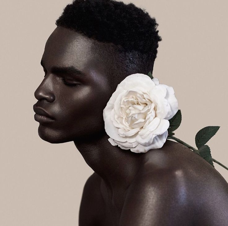 a closeup portrait of a handsome black man with a white rose behind his ear. his skin is smooth and dark, and he is looking away from the camera with a soft expression on his face. the image is a study in contrasts, with the light skin of the rose against the dark skin of the man. the flower is a symbol of beauty and elegance, while the man's dark skin represents power and strength.