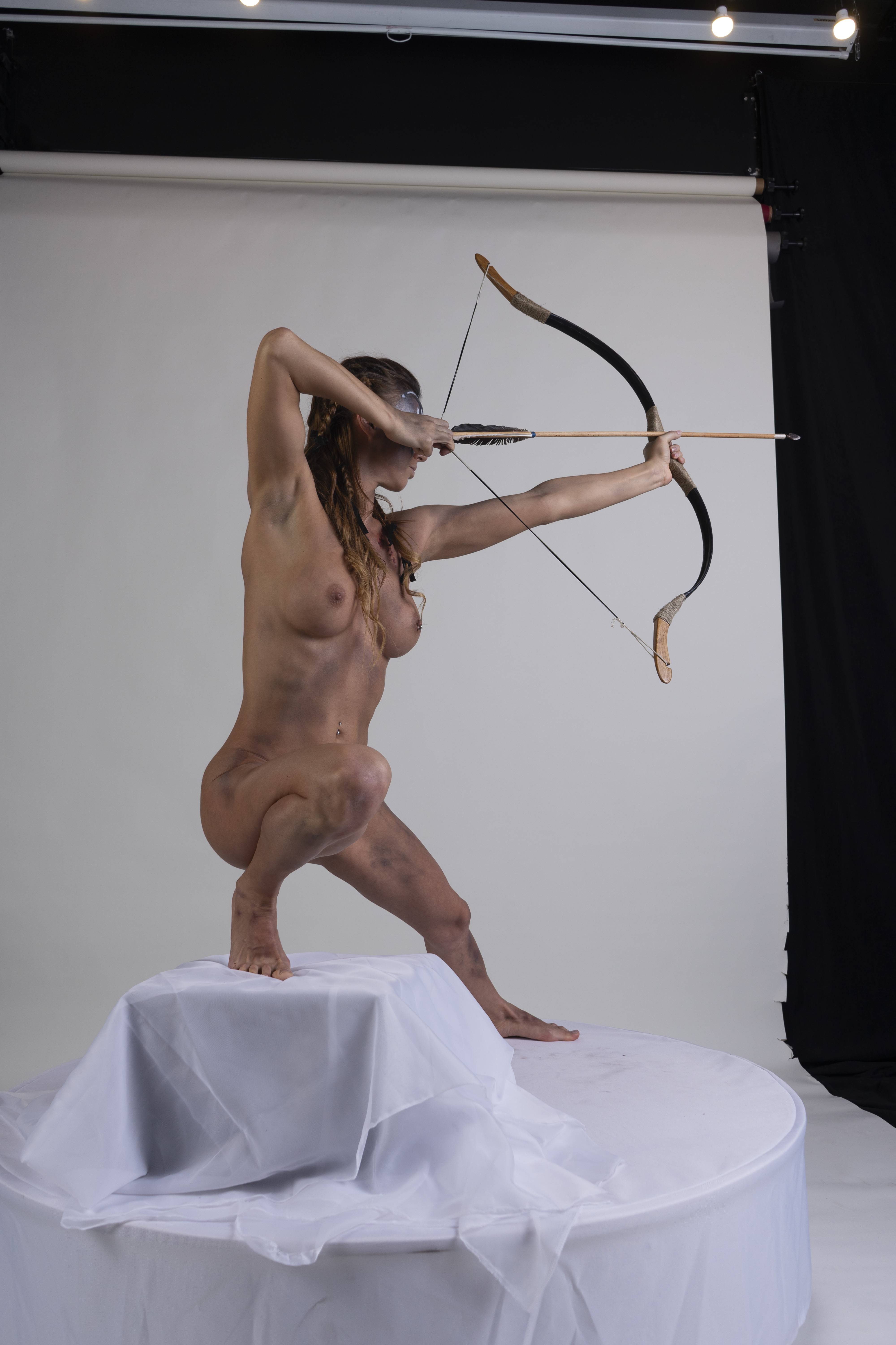 a nude, muscular woman in a warrior pose is positioned on a white draped platform in a studio setting. she is holding a bow and arrow, aiming at a target out of frame. she has tribal markings on her face and body, suggesting a mythical or warrior theme. the backdrop consists of a white wall and a black curtain.