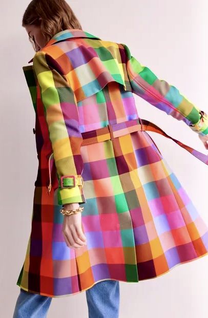 a woman is shown from the back, wearing a bright, colorful plaid trench coat and blue jeans. the trench coat is a bold, eyecatching pattern with a mix of different colors, including red, orange, yellow, green, purple, and brown. it has a belted waist and reaches about midcalf. the woman has short brown hair and is wearing a gold bracelet on her left wrist. the background is a plain, light color.