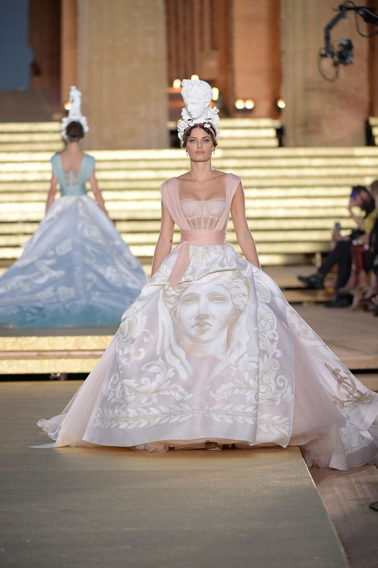 a model walks the runway in a white gown with pink floral accents and a white headpiece. the gown is adorned with a large floral design and a  face of a woman printed on the front. she is wearing her hair pulled back and her make up is natural. the background is a  stage with an audience seated in the back.
