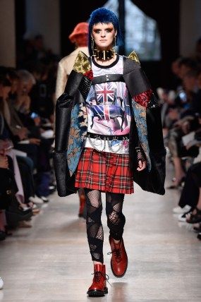 a female model walks down a runway in a black leather jacket, red and black plaid skirt, and red boots. she has blue hair and wears a variety of accessories including sunglasses, a black choker, and a silver belt. the model is walking in front of a blurred background of other people.
