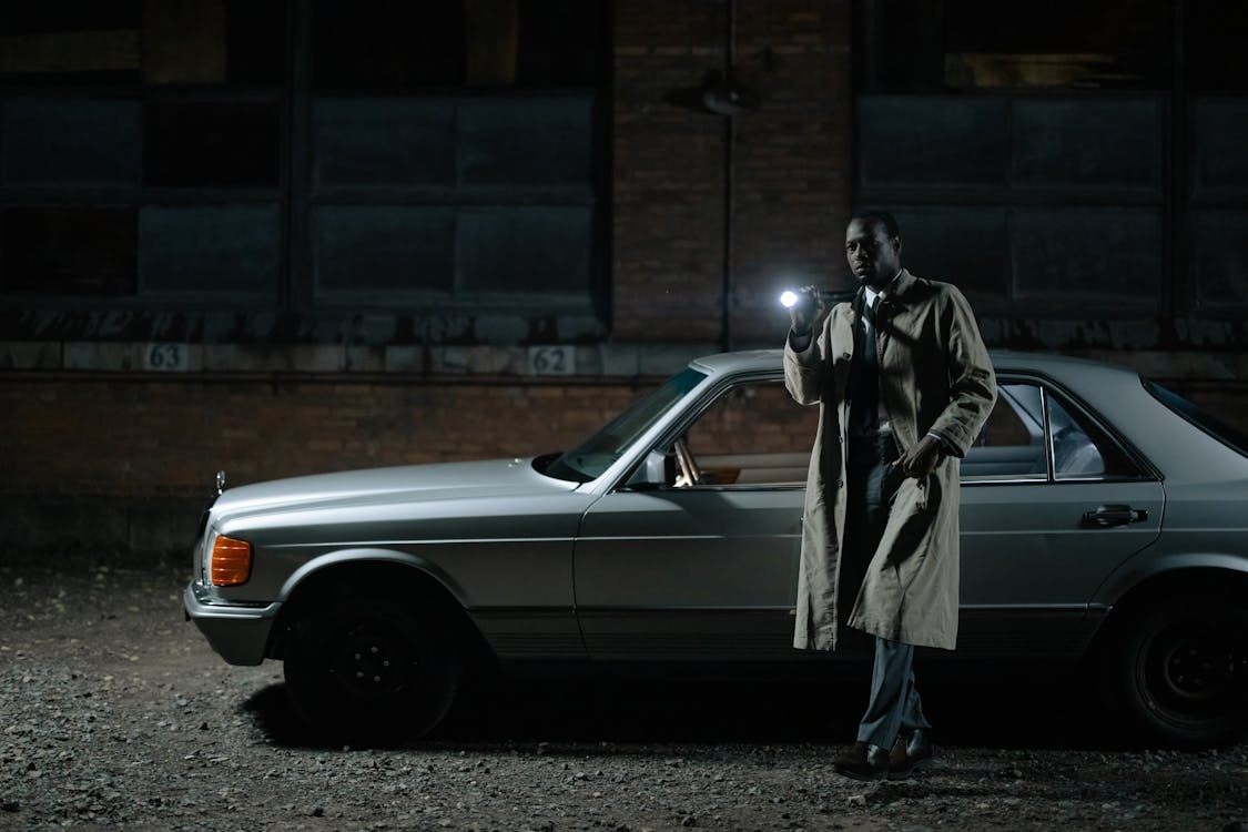 a man in a trench coat stands beside a vintage silver mercedesbenz at night, holding a flashlight. he appears to be a detective, possibly investigating a crime scene. the setting is urban and dark, creating a moody and atmospheric scene reminiscent of film noir.  the car is parked against a brick wall in a dimly lit alley or street.