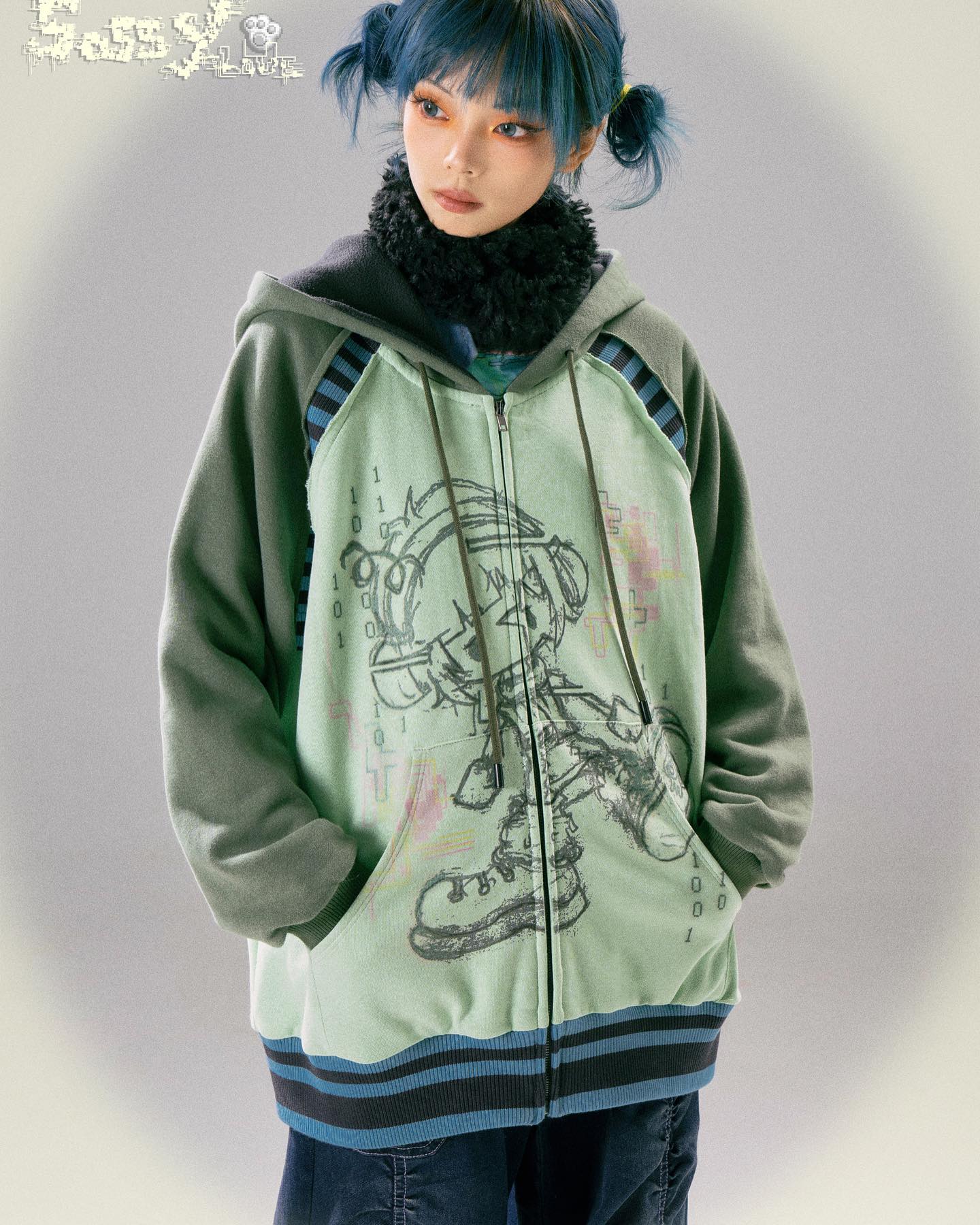 a closeup shot of a young woman modeling a pastel green zipup hoodie. the hoodie features a cartoon graphic of an animestyle character and teal and dark teal stripes along the sleeves and bottom hem. the model has short, teal hair styled with side bangs and two small pigtails on either side of her face. she is wearing the hoodie with darkwash jeans and a faux fur collar underneath. the background is a muted, light gray, which focuses attention on the garment. the image displays a  streetwear fashion style that is reminiscent of  kawaii and y2k aesthetics.