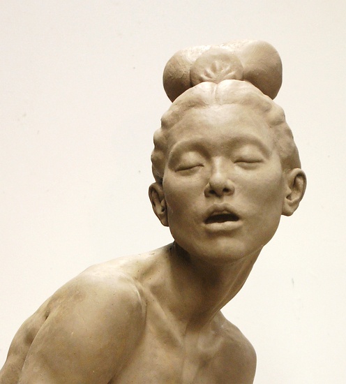 a clay sculpture of a woman's bust with her eyes closed and mouth slightly open. she has a bun on top of her head. the sculpture is set against a plain background.