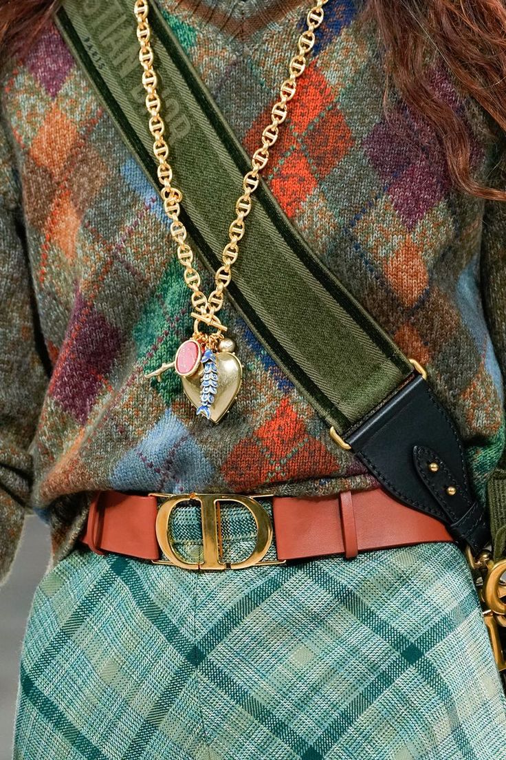 closeup shot of a woman's outfit. she is wearing a multicolored, patterned sweater, a teal plaid skirt, a brown leather belt with a gold dior buckle, and a gold necklace with charms. a green and black shoulder bag is slung across her body. the overall style is autumnal and sophisticated, with a mix of textures and colors.  the focus is on the details of the clothing and accessories, showcasing the highend, designer aesthetic.