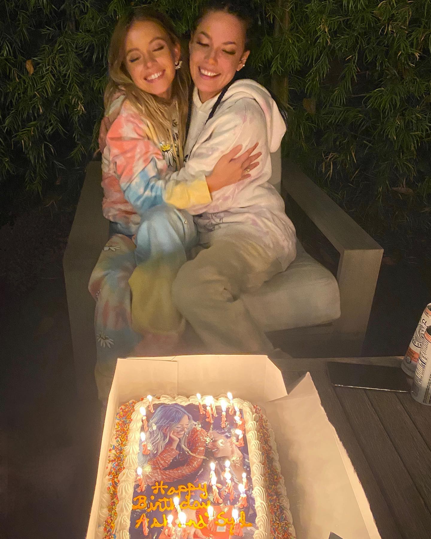 the image shows ashley tisdale and sarah hyland hugging each other.  they are sitting on a patio at night. ashley is wearing a pastel tiedye sweatshirt and pants, while sarah is in a cream tiedye hoodie and pants. between them, on a table, sits a birthday cake with a photo of ashley tisdale on it.  the cake is lit with candles. the overall atmosphere is warm and celebratory, suggesting a birthday party for ashley tisdale.